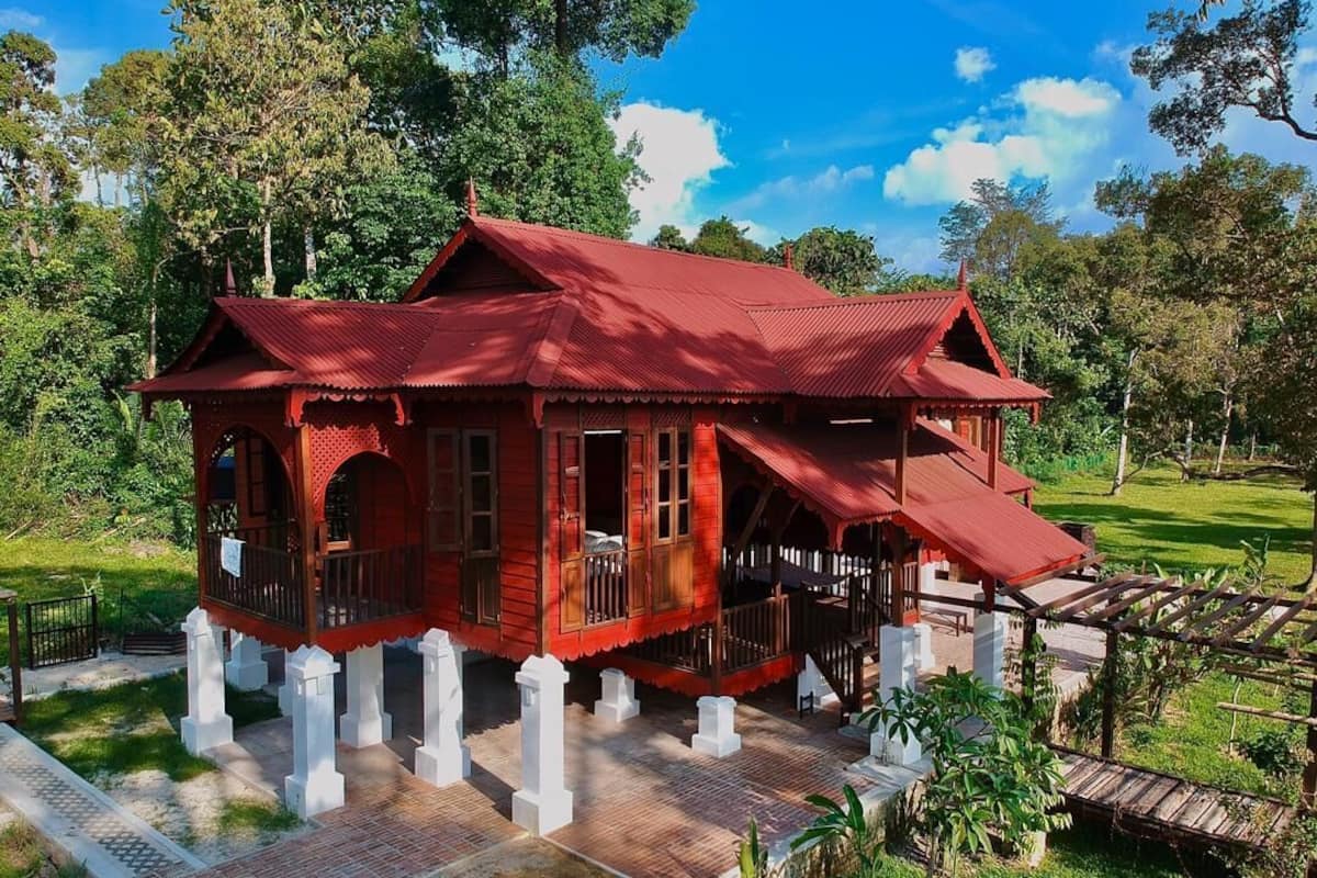 Une maison traditionnelle en bois, peinte en rouge, dotée d’un balcon et entourée d’une végétation luxuriante et de piliers blancs.