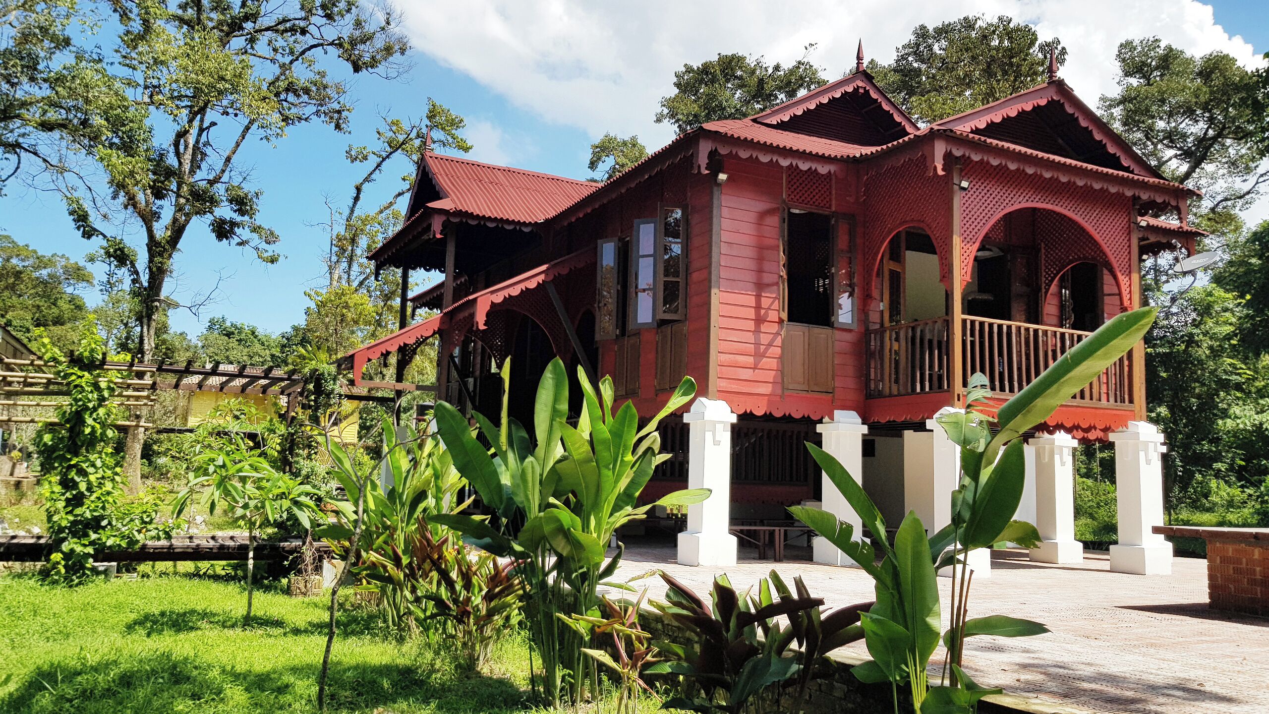 A traditional red wooden house with a balcony, surrounded by lush greenery and trees.