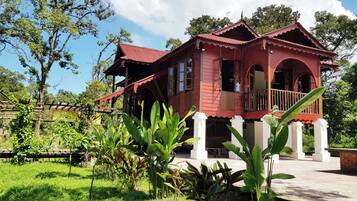 A traditional red wooden house with a balcony, surrounded by lush greenery and trees.