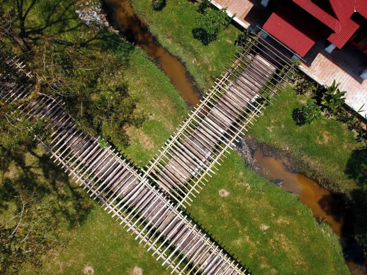 An aerial view of a wooden bridge over a stream, surrounded by greenery and a building with a red roof.