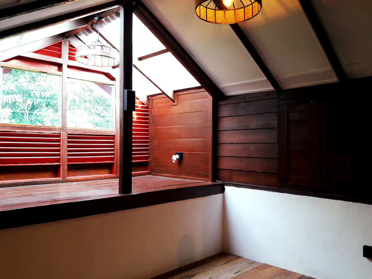 Una habitación con techo inclinado, paredes de madera y ventana con persianas rojas.