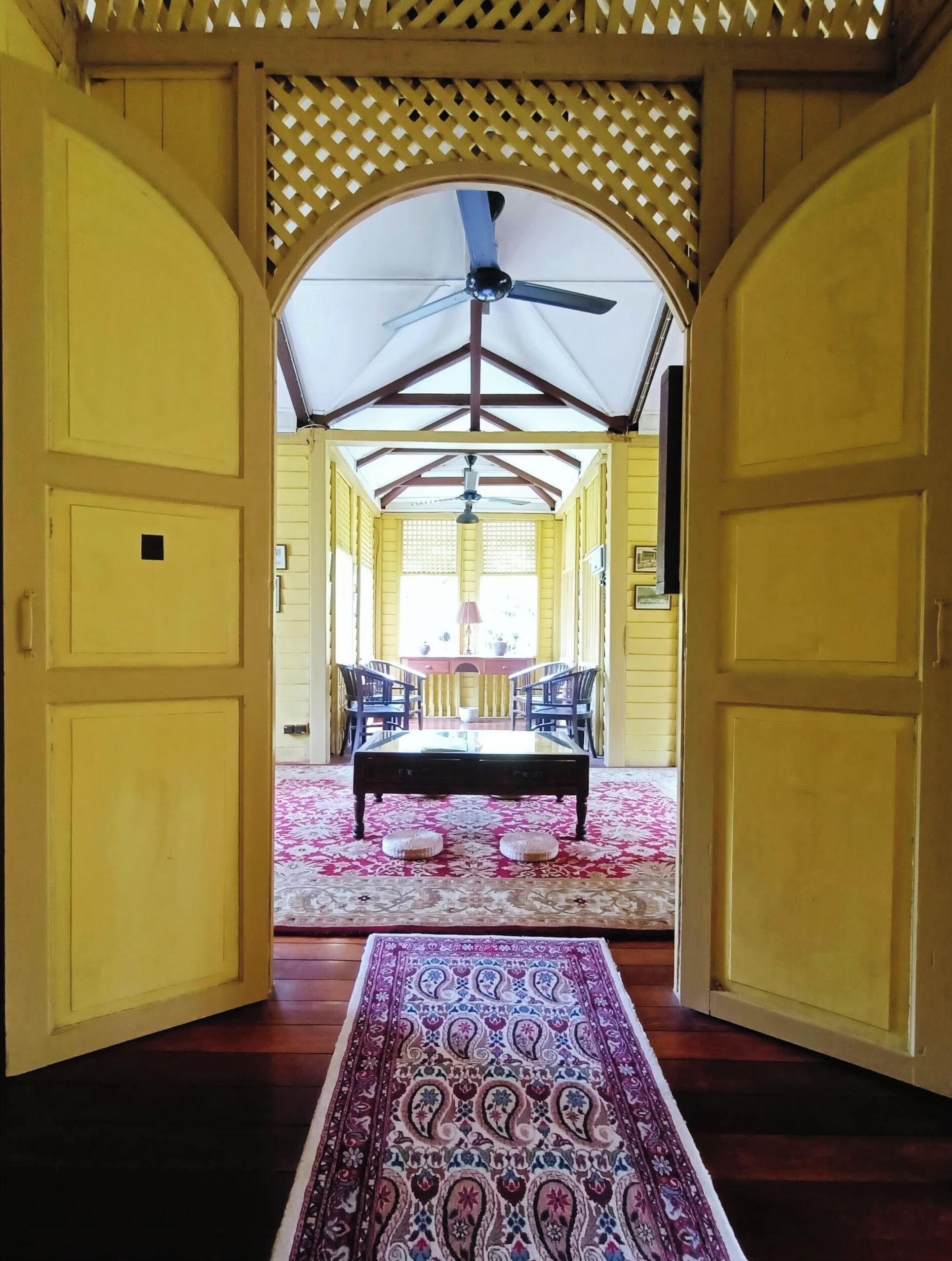 Quarto com piso de madeira, tapete estampado, ventilador de teto e mesa com cadeiras.