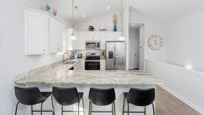 Una cocina moderna con armarios blancos, una isla de mármol, taburetes negros de barra, electrodomésticos de acero inoxidable e iluminación colgante.