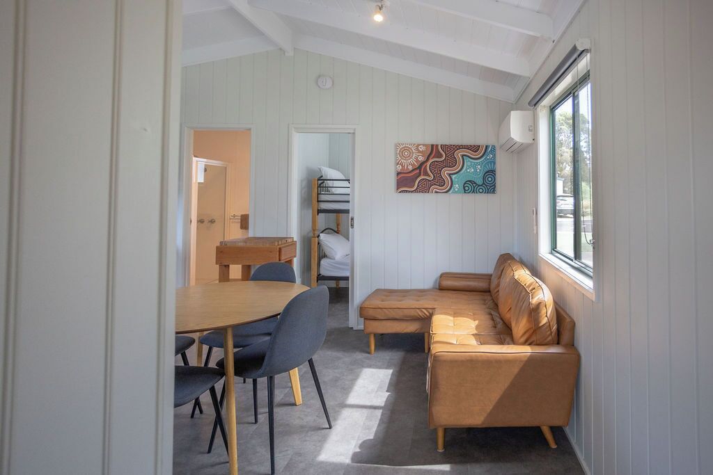 A cozy room with a wooden dining table, two blue chairs, a brown leather sofa, and a colorful wall art piece.