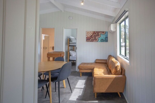 A cozy room with a wooden dining table, two blue chairs, a brown leather sofa, and a colorful wall art piece.