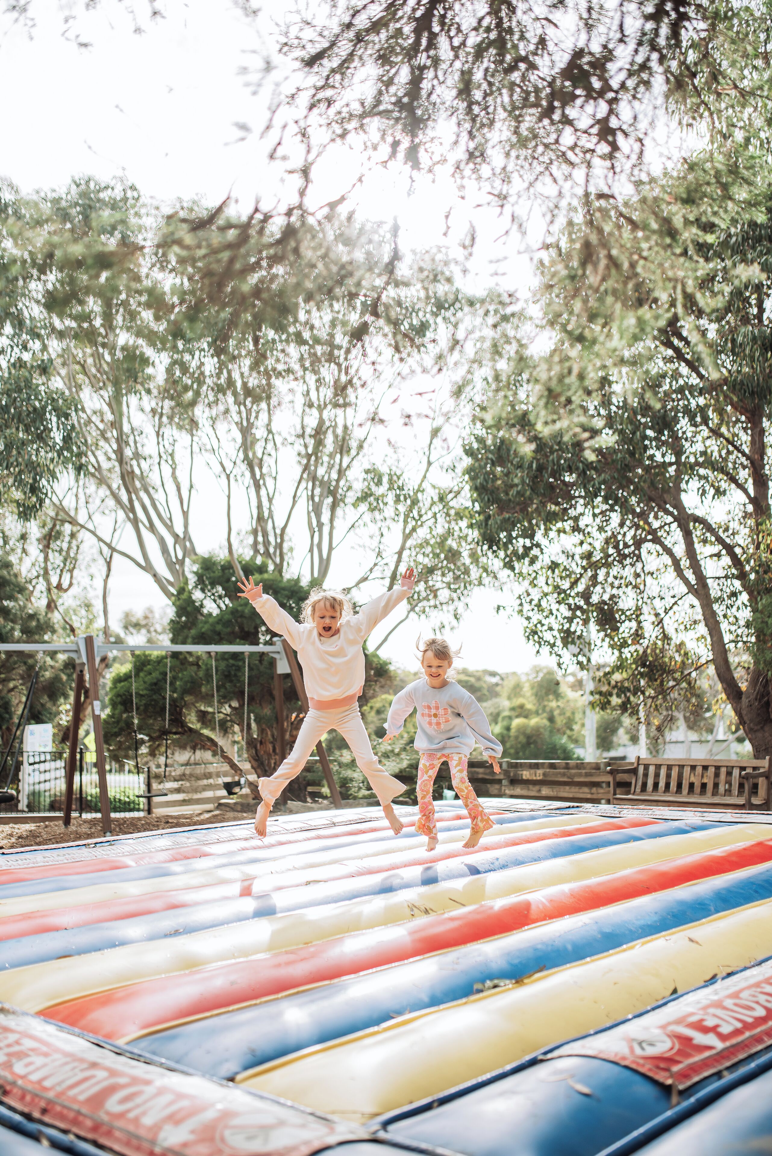 Two children jumping on a colorful inflatable raft in an outdoor setting with trees and a fence in the background.