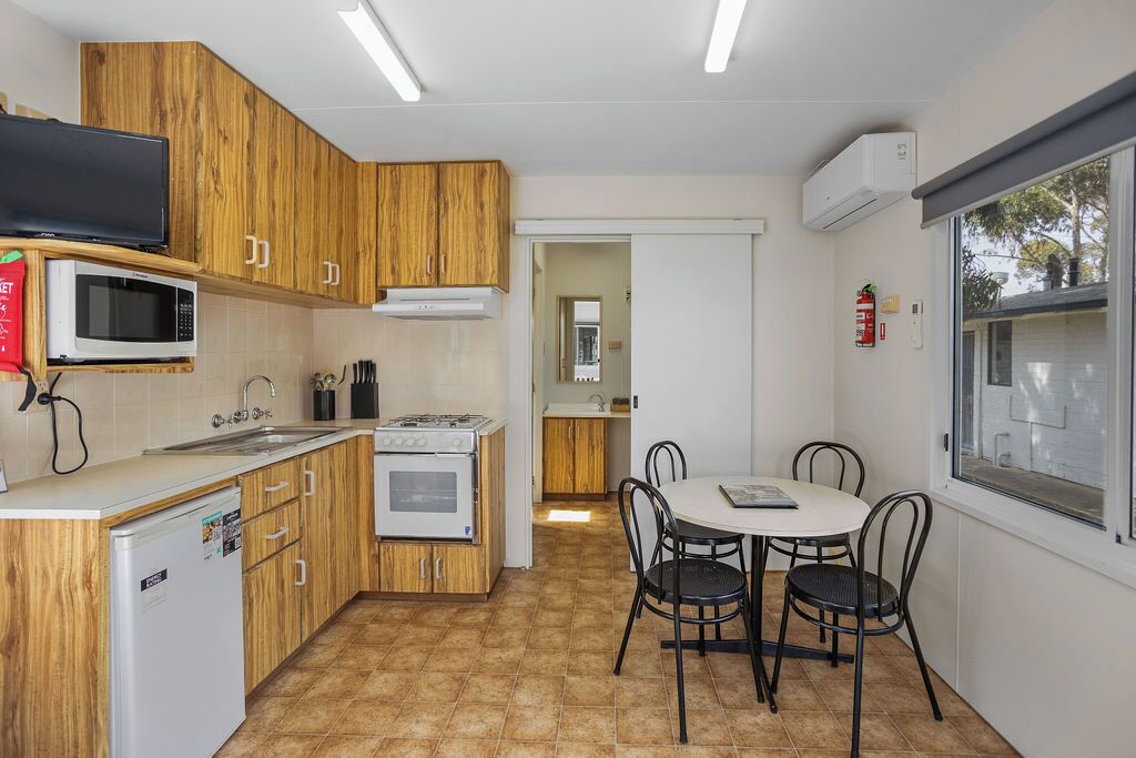 Una cocina funcional con armarios de madera, microondas, estufa y lavamanos. Hay una mesa de comedor con sillas y una ventana con persianas.