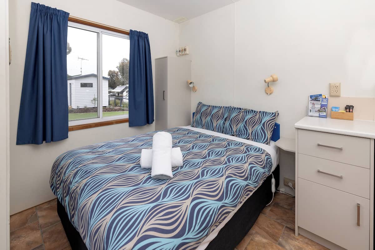 Ein Schlafzimmer mit Bett, einem Fenster mit Vorhängen, einem Nachttisch mit Pflegeprodukten und Blick auf ein Haus.