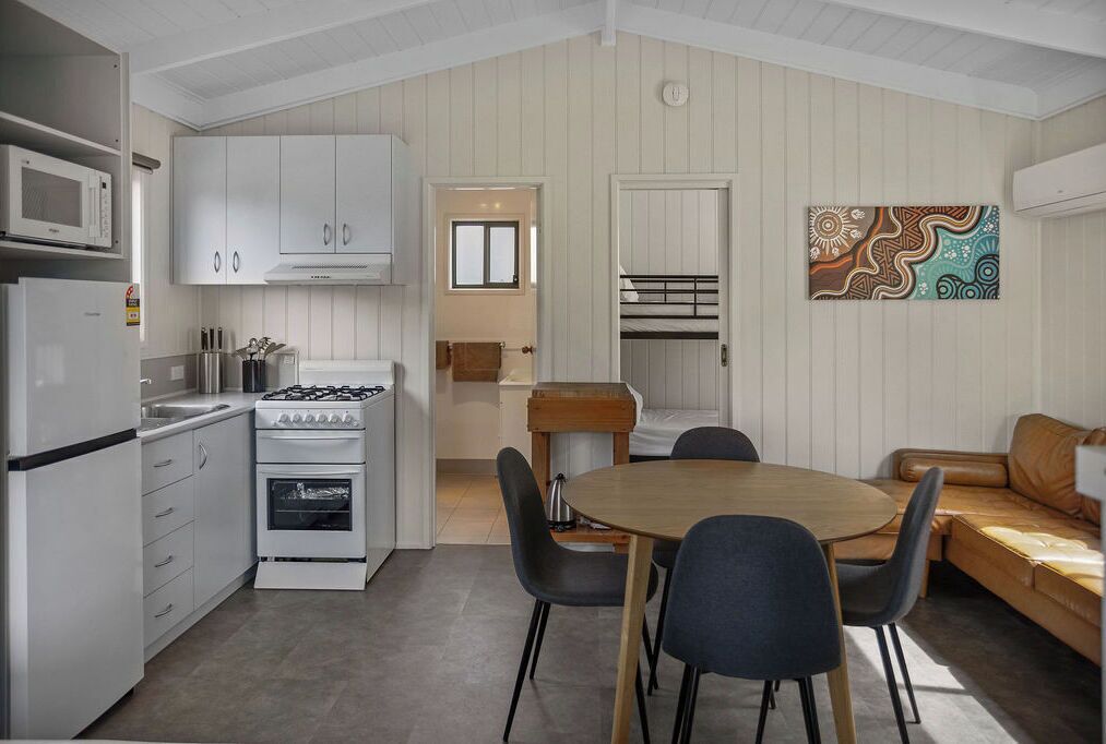 Een kleine keuken met witte kastjes, een gasfornuis en een magnetron. Er is een eethoek met een houten tafel en stoelen, en een bruine leren bank.