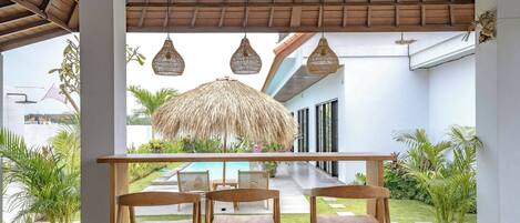 An outdoor seating area with wooden chairs and a thatched umbrella, overlooking a pool and garden.
