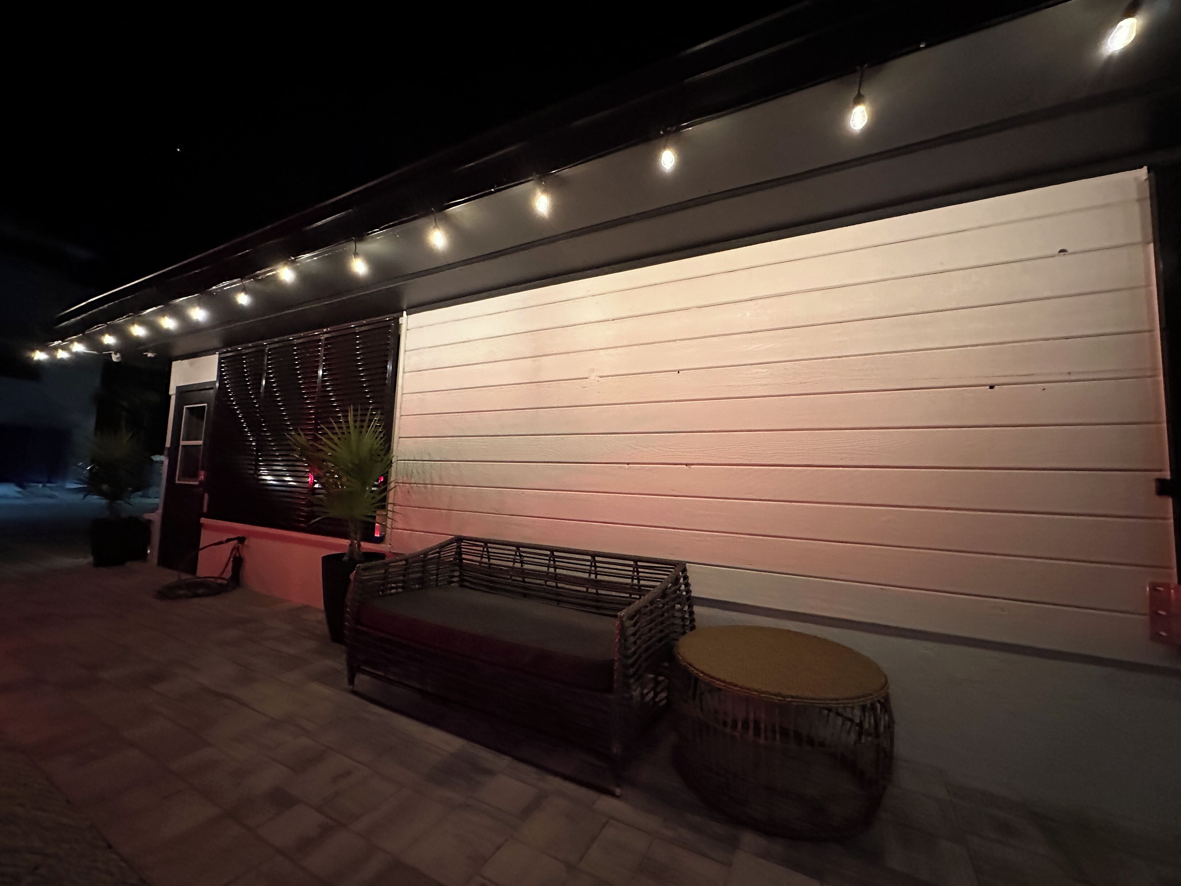 Un espacio al aire libre con un banco, una mesa redonda, una planta en maceta y luces colgantes a lo largo de la pared.