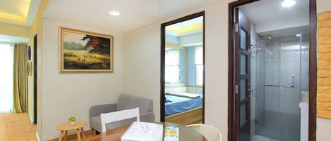 A hotel room with a wooden dining table, a small round table with a plant, a grey sofa, a bed with a blue bedspread, and a glass-enclosed shower area.