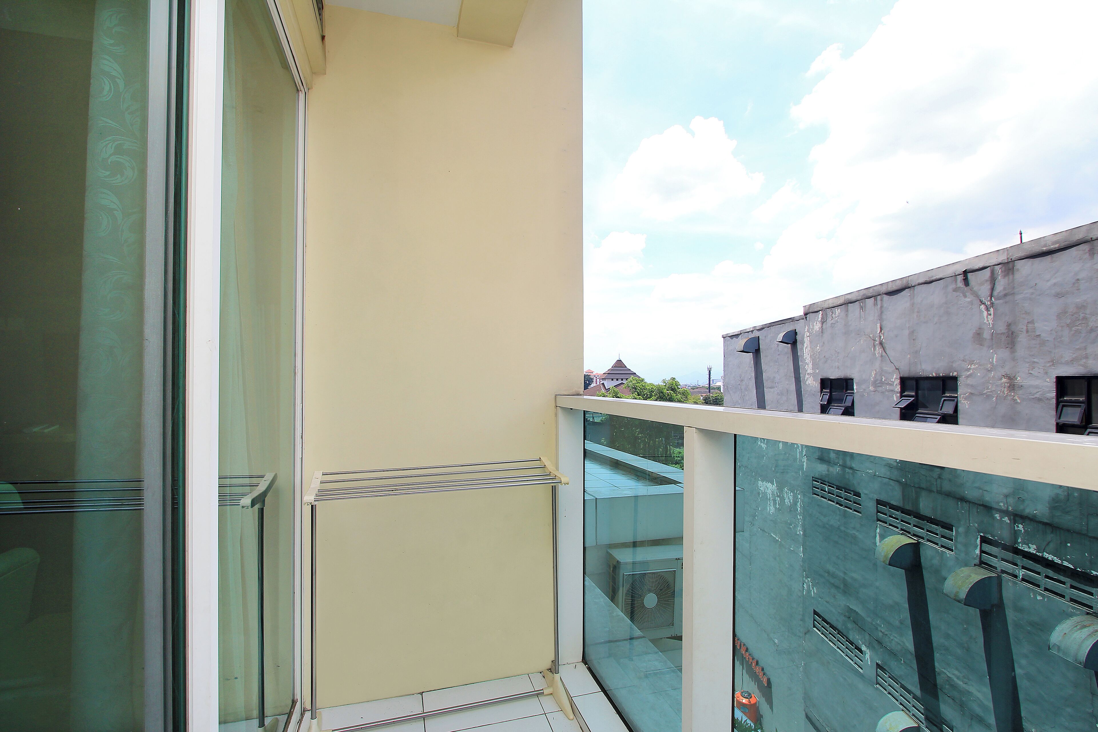 Un balcón con puerta de cristal, vista de un edificio con unidades de aire acondicionado visibles, y cielo despejado con nubes.