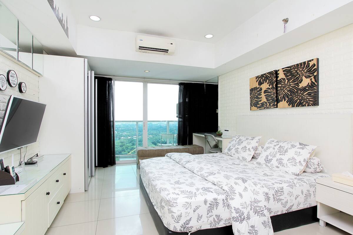 A modern bedroom with a large bed, a desk with a computer, a TV, and a balcony with a city view.