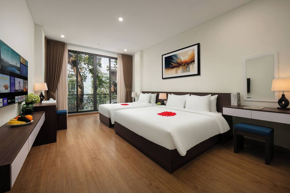 A hotel room with two beds, a desk, a chair, a TV, and a balcony with a view of trees.