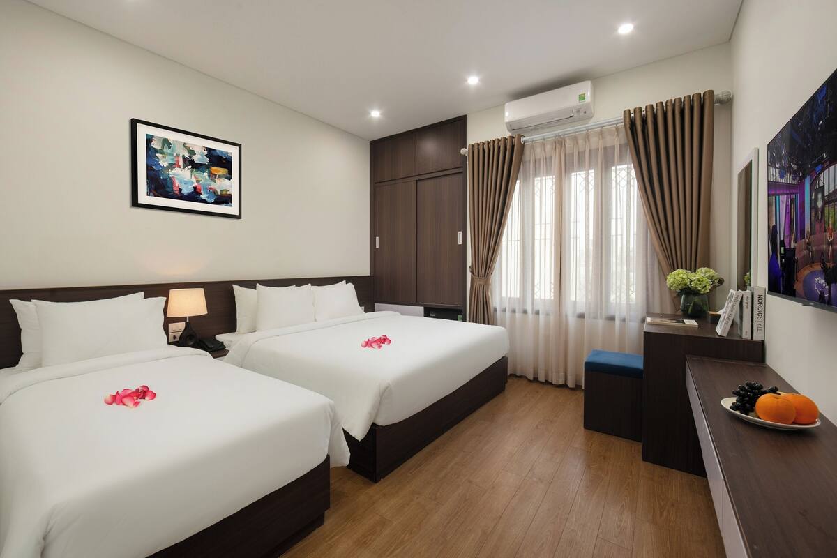 A hotel room with two beds, a desk with a plate of fruit, a painting on the wall, and an air conditioning unit.