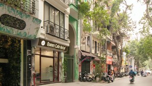 A street view with the Queen Cafe Hotel, featuring a storefront with large glass windows, a balcony with wrought iron railings, and a green awning. - Queen Cafe Hotel (Hanoi)