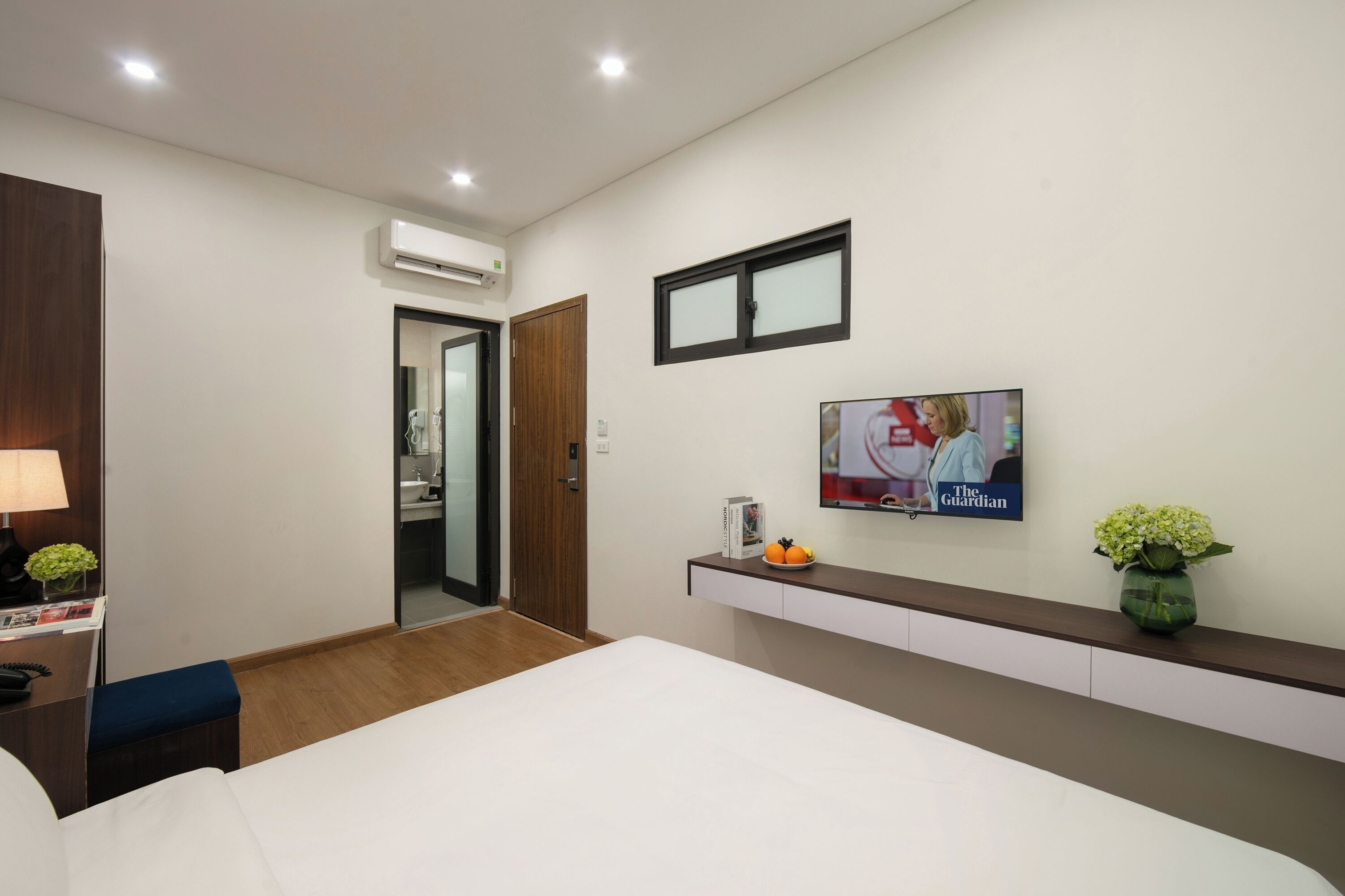 A hotel room with a bed, a desk with a lamp, a TV on the wall, and a small shelf with a vase and a book.