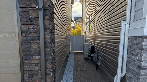 A narrow alleyway with a stone wall on one side and a building with horizontal siding on the other. There is a metal gate in the center, and a utility box is visible on the right side. - Cozy 2BR Suite | Near CTrain, Spruce Meadows & Parks | Modern Comforts Await! (Calgary)