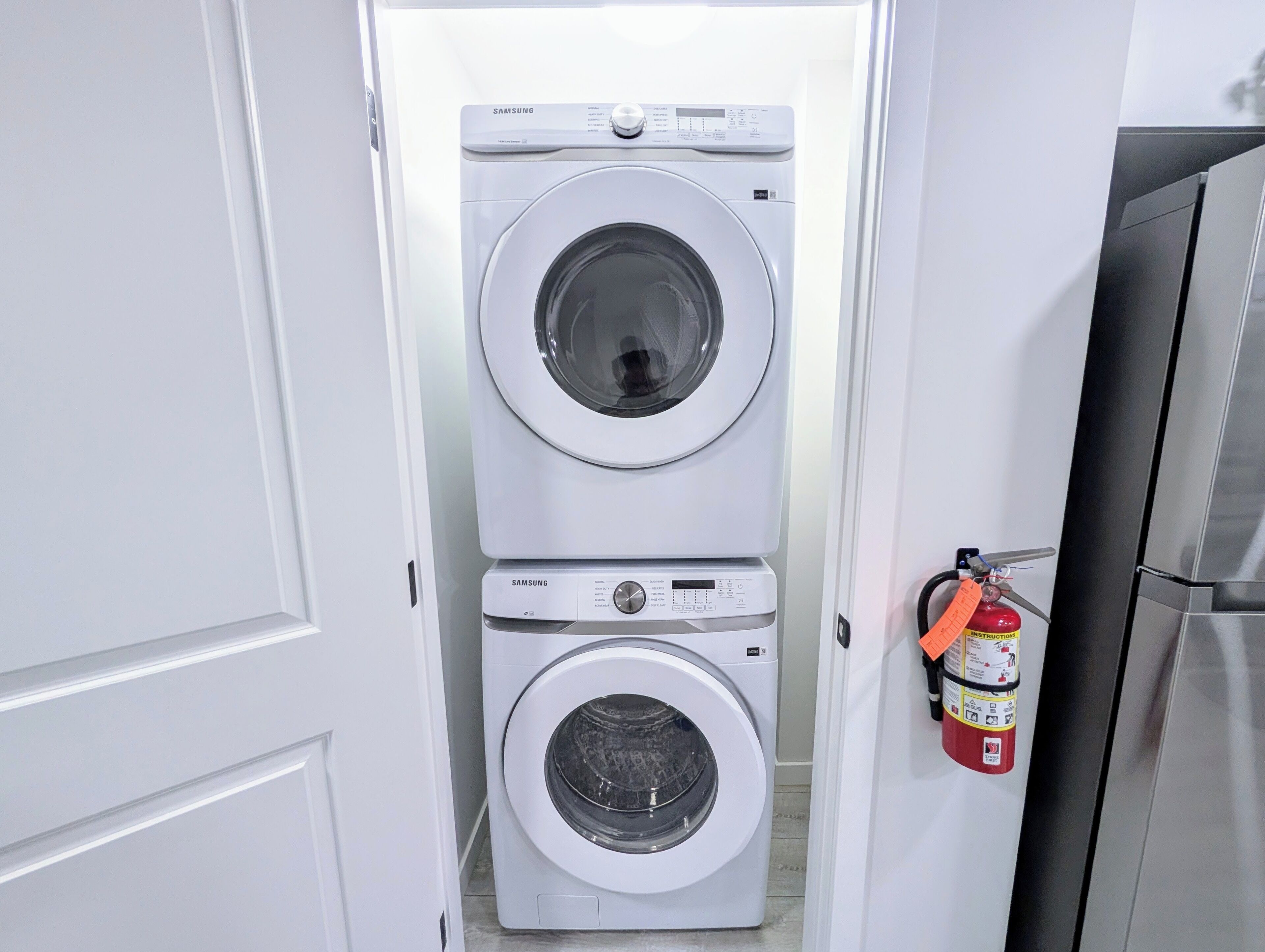 A laundry room with two stacked Samsung washing machines and a fire extinguisher mounted on the wall.