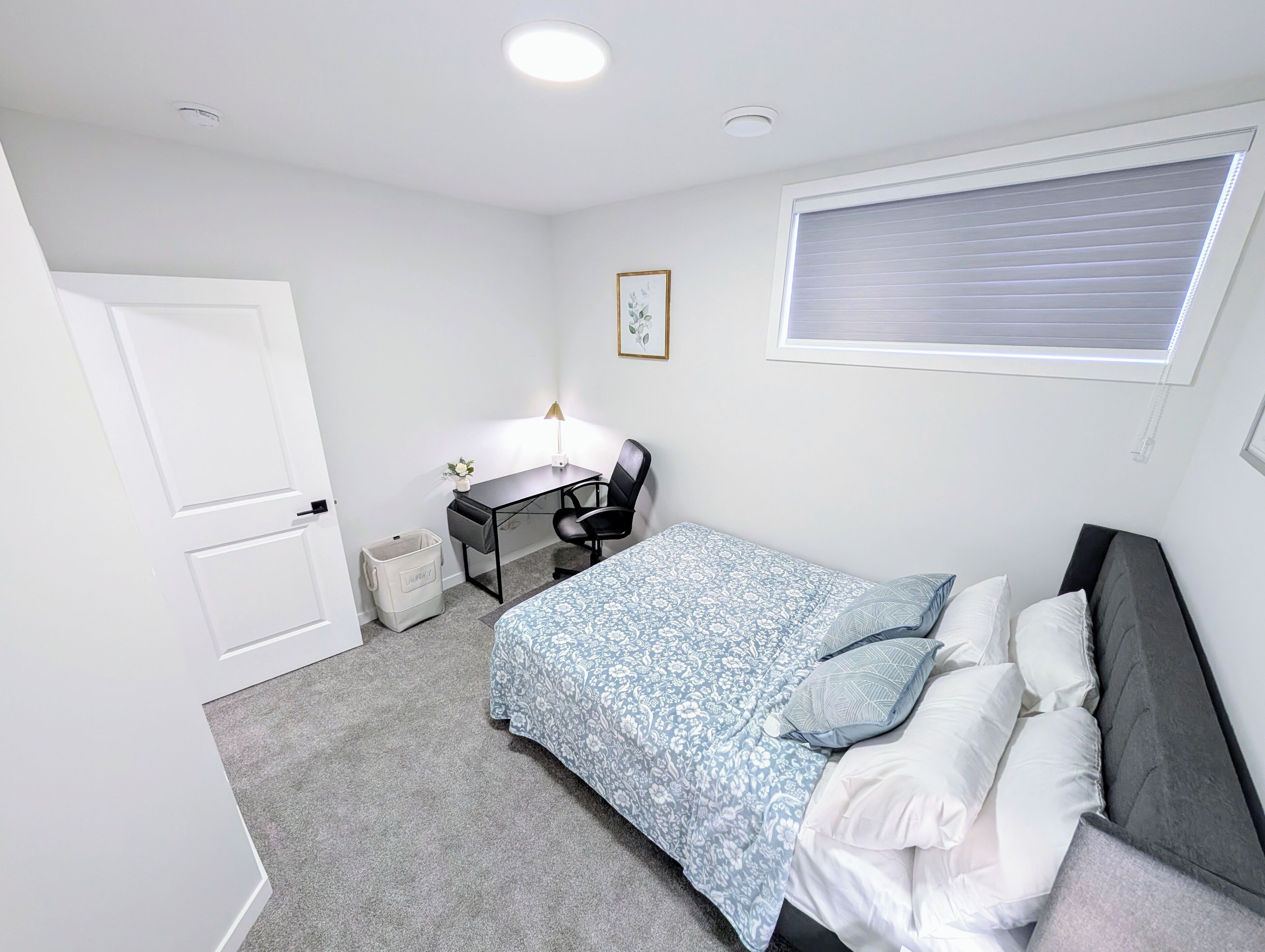A bedroom with a bed, a desk, a chair, a window with blinds, and a door.