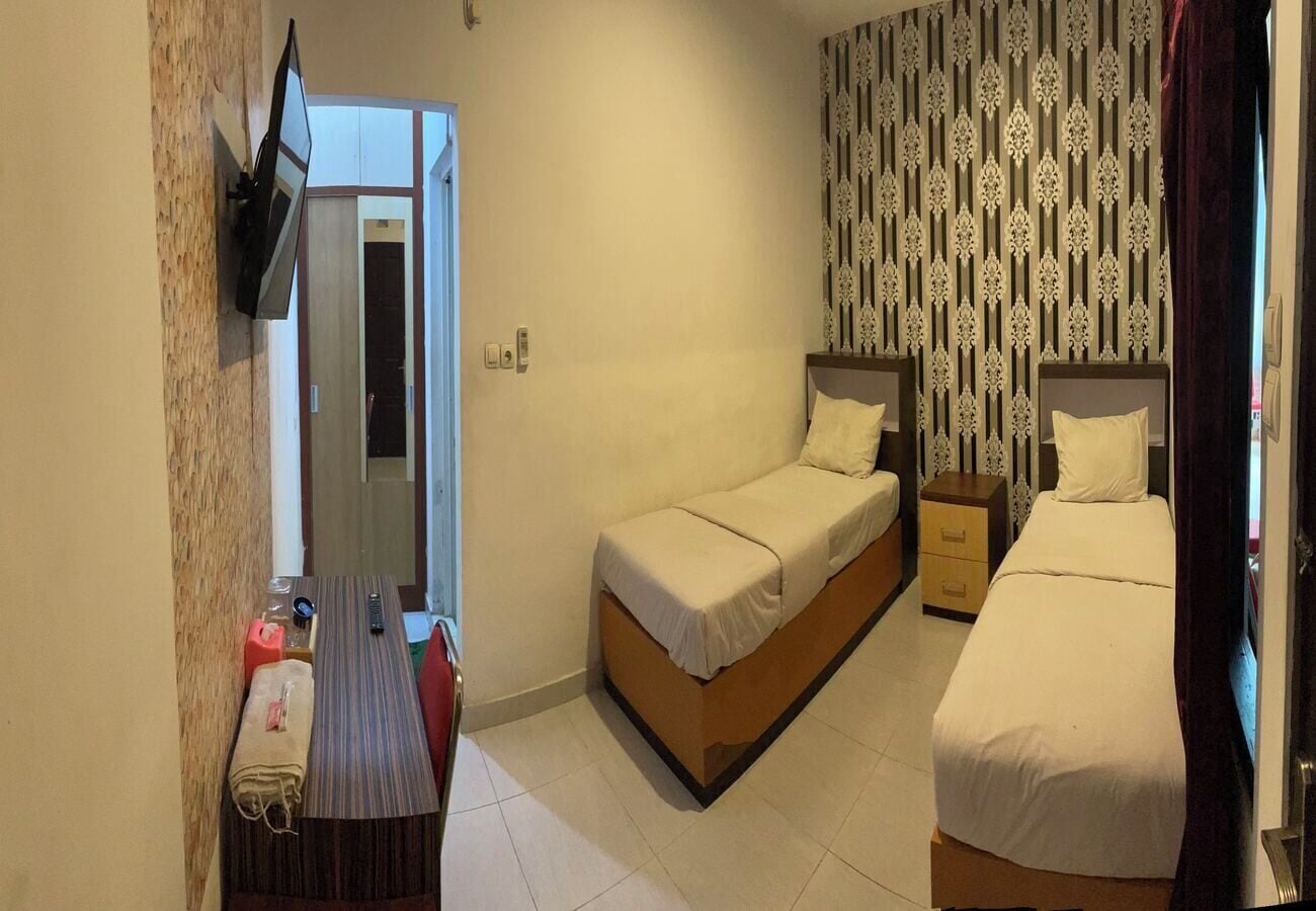 A small hotel room with two single beds, a wooden nightstand, a small table with a chair, a TV mounted on the wall, and a patterned wallpaper.