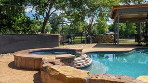 A swimming pool with a circular hot tub, surrounded by rocks and a covered pavilion. - Pool, Spa, Bar + Game Room - Huntsville Lake House (Huntsville)