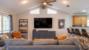 A living room with a gray sectional sofa, a flat-screen TV, a ceiling fan, and a dining area with bar stools. - Pool, Spa, Bar + Game Room - Huntsville Lake House (Huntsville)