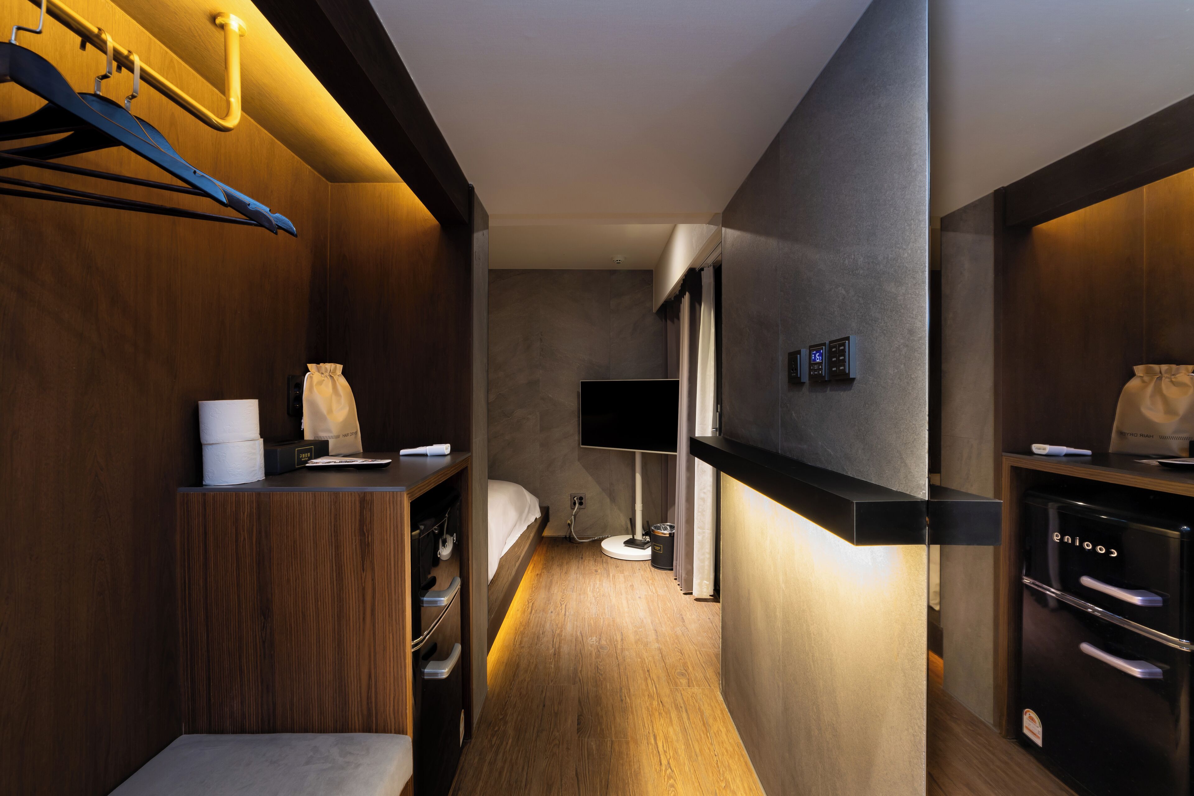 Habitación de hotel pequeña con piso de madera, cama, escritorio, armario, televisor integrado y minibar.