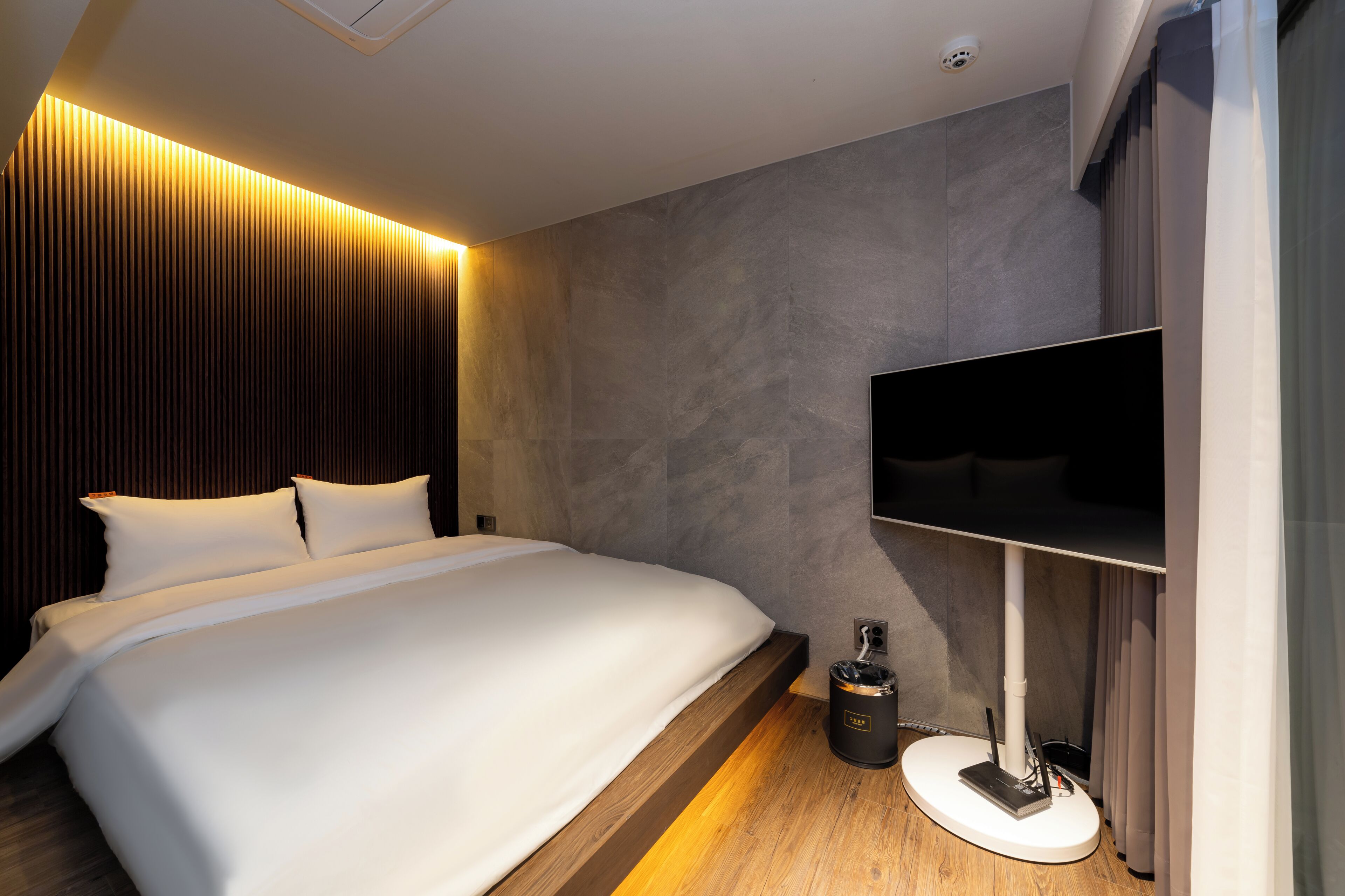 A modern hotel room with a large bed, a flat-screen TV mounted on the wall, and a small table with a trash bin.