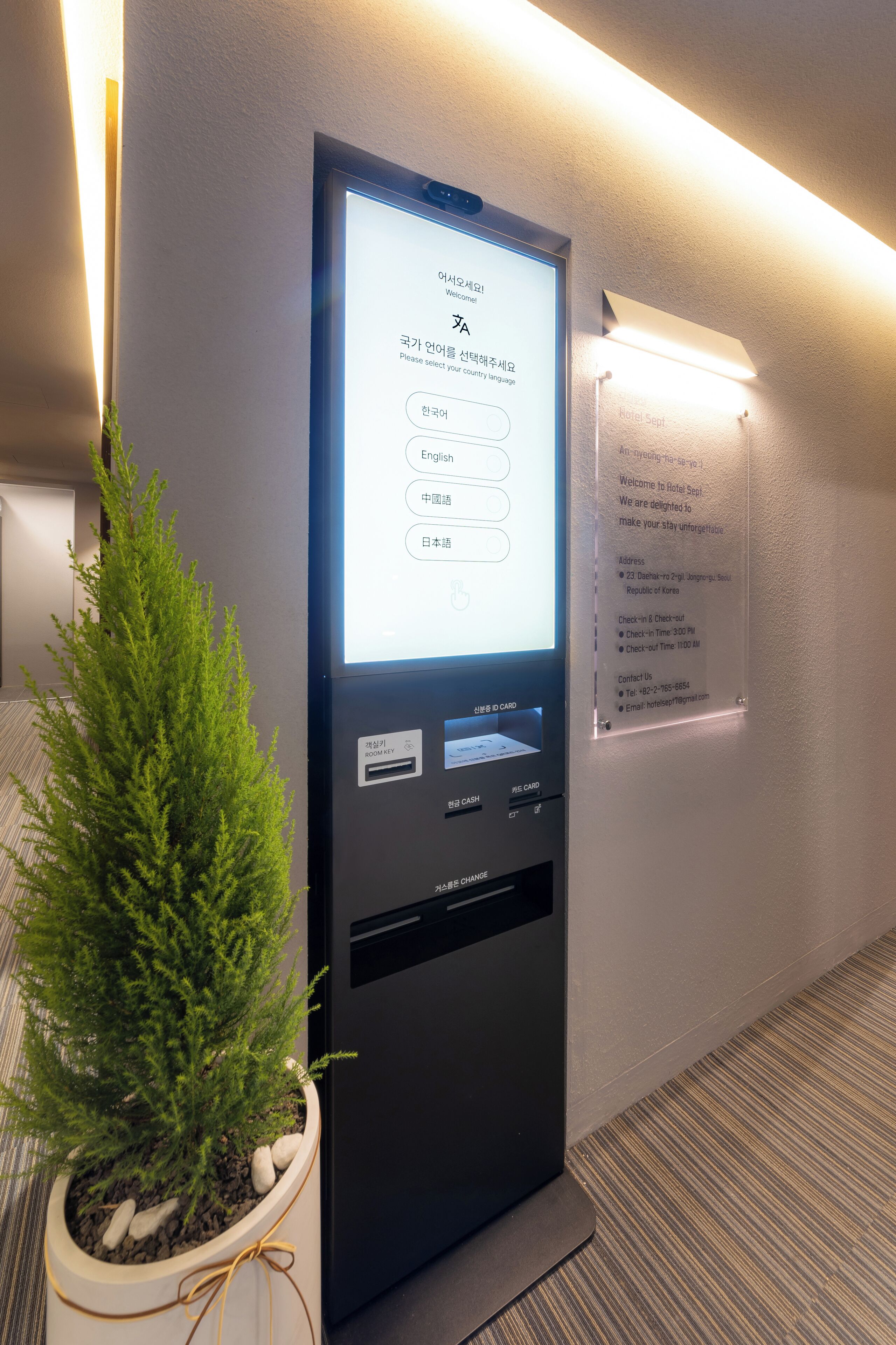 Un quiosco digital de registro de entrada con una pantalla que muestra un menú de selección de idioma, una pequeña planta en maceta a un lado y un letrero de bienvenida en la pared.