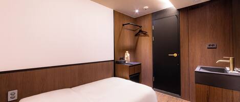 A hotel room with a bed, a wooden wardrobe, a black door, a small bedside table with a bottle, and a sink with a gold faucet.