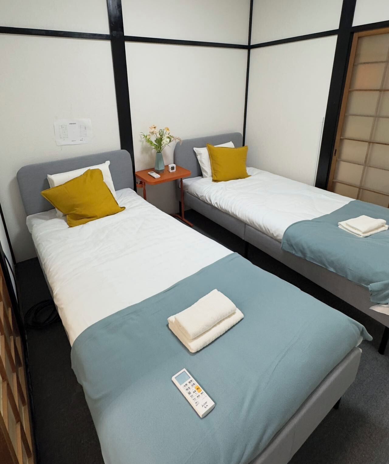 A hotel room with two single beds, each with a grey headboard, white bedding, and a mustard-colored pillow. There is a small wooden nightstand with a vase of flowers and a remote control. A folded white towel is placed on the bed in the foreground.