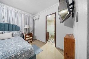 A bedroom with a bed, a wooden nightstand, a TV mounted on the wall, and a bathroom visible through an open door. - Cozy 2-bedroom Apartment in Santo Domingo's Heart (Santo Domingo)