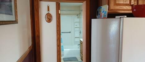Eine Küche mit einem weißen Kühlschrank, Holzschränken und einer Tür zu einem Badezimmer mit Badewanne und Dusche.