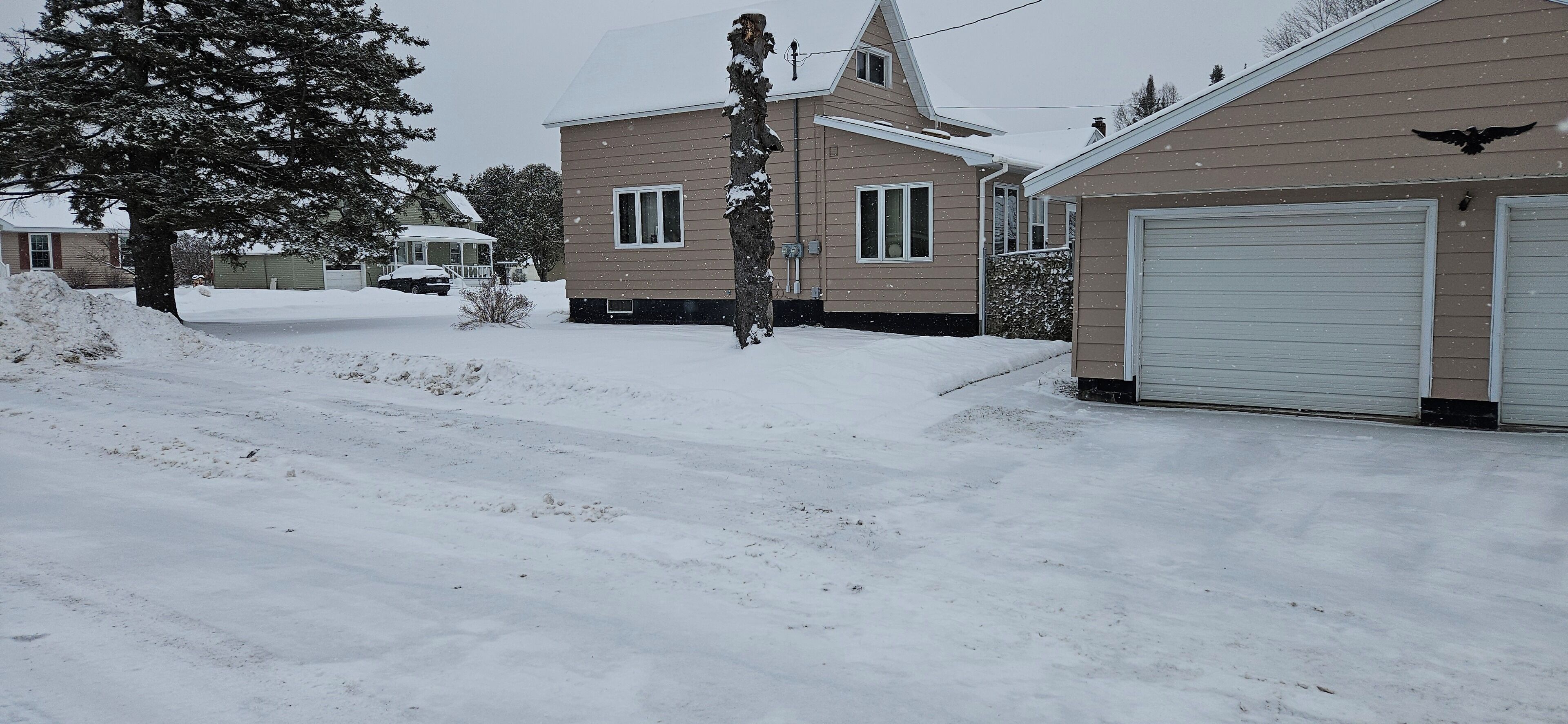 A residential area with a house and garage, surrounded by snow-covered ground and trees.