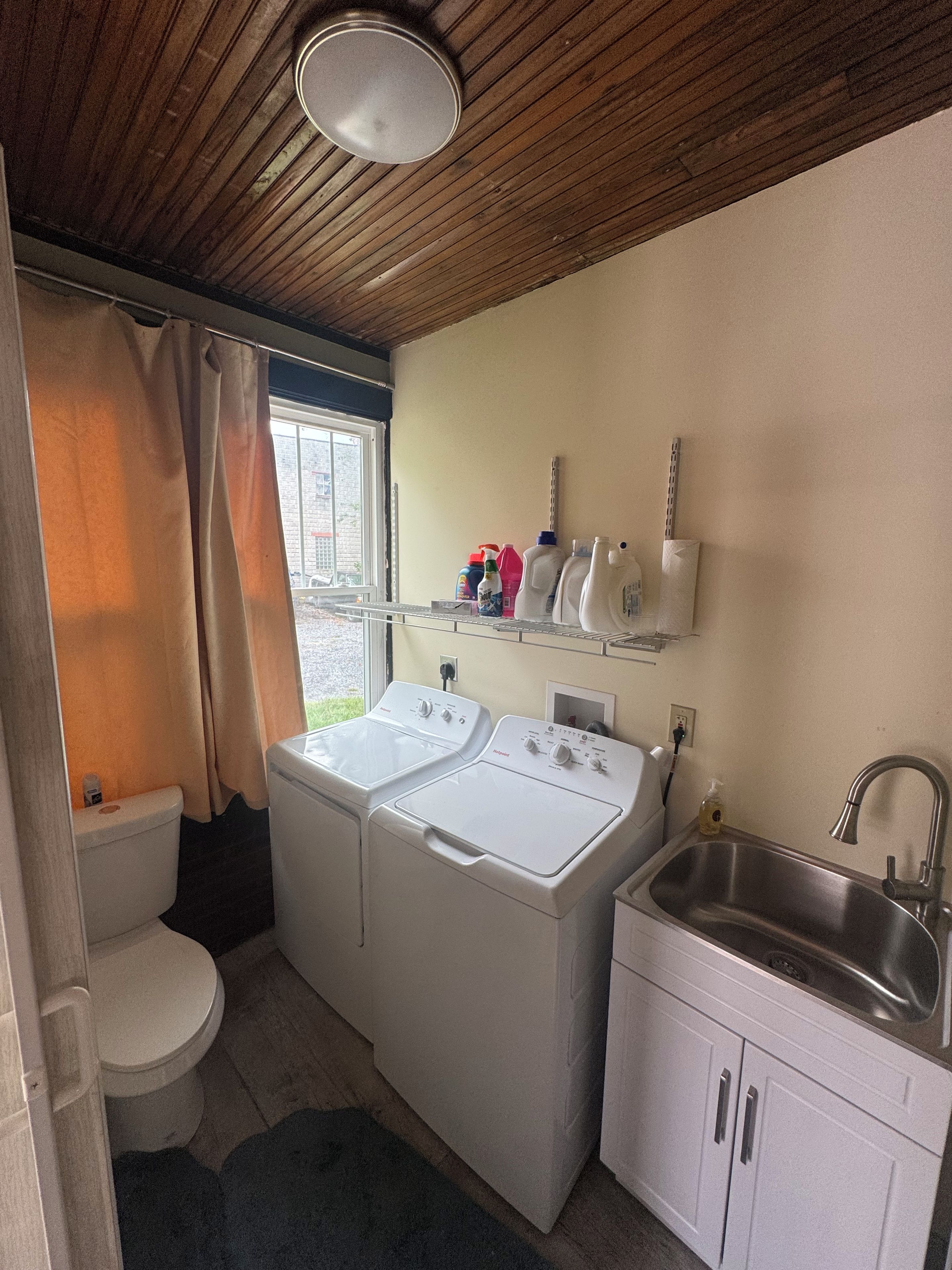 A compact laundry room with two front-loading washing machines, a sink, and a window with curtains.