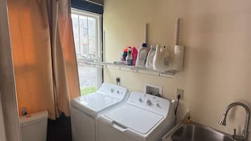A compact laundry room with two front-loading washing machines, a sink, and a window with curtains.