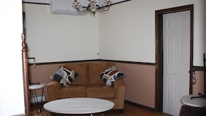 A corner room with a brown sofa, two patterned pillows, a round table, a small stool, a white door, and a chandelier. - All one floor Charming large2 bedroom, LR, sunrm, kitchen, 1 Full & 1 half bath (Roaring Spring)