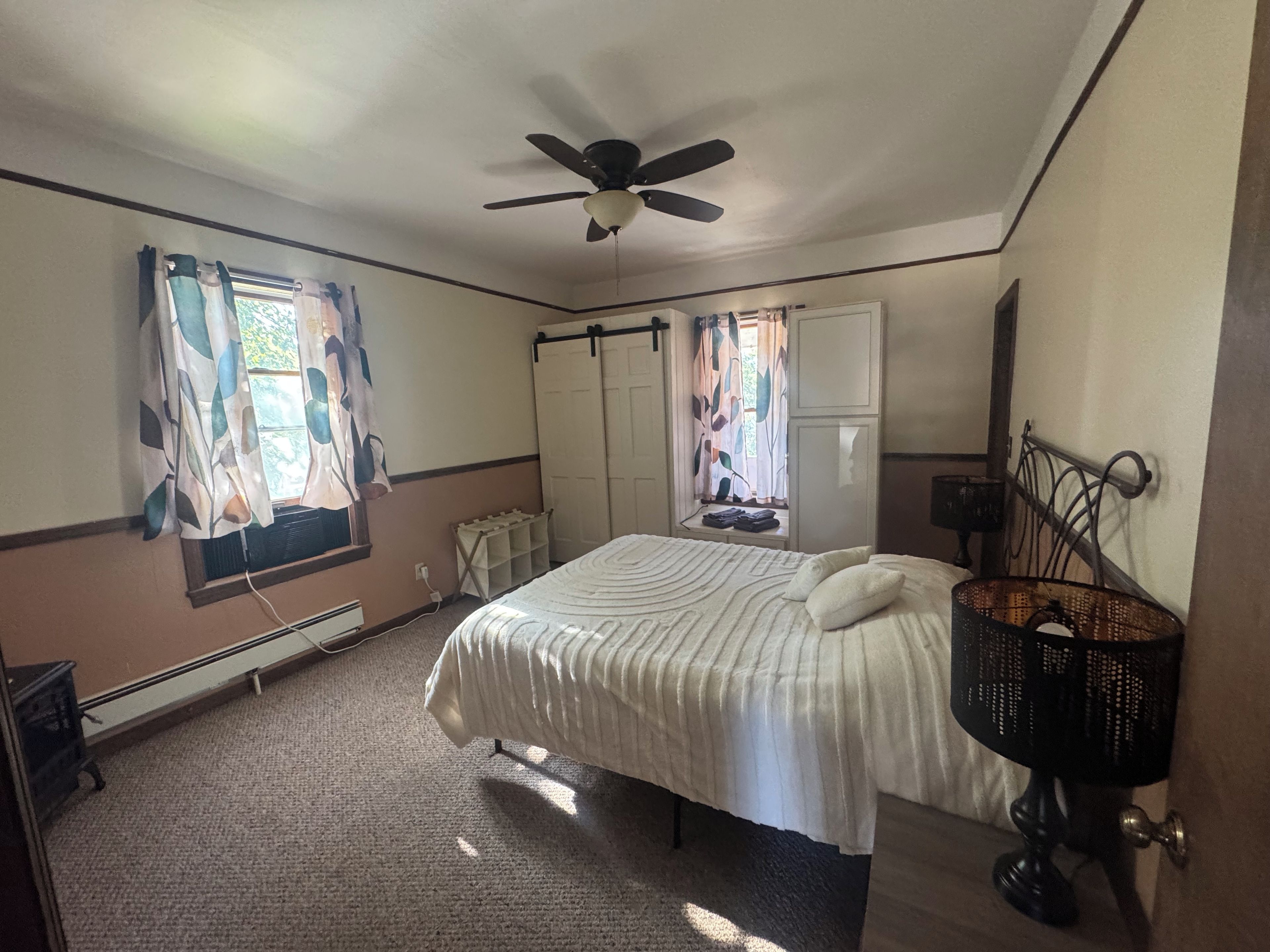 Une chambre à coucher avec un lit, un ventilateur de plafond, un radiateur et une fenêtre avec des rideaux.