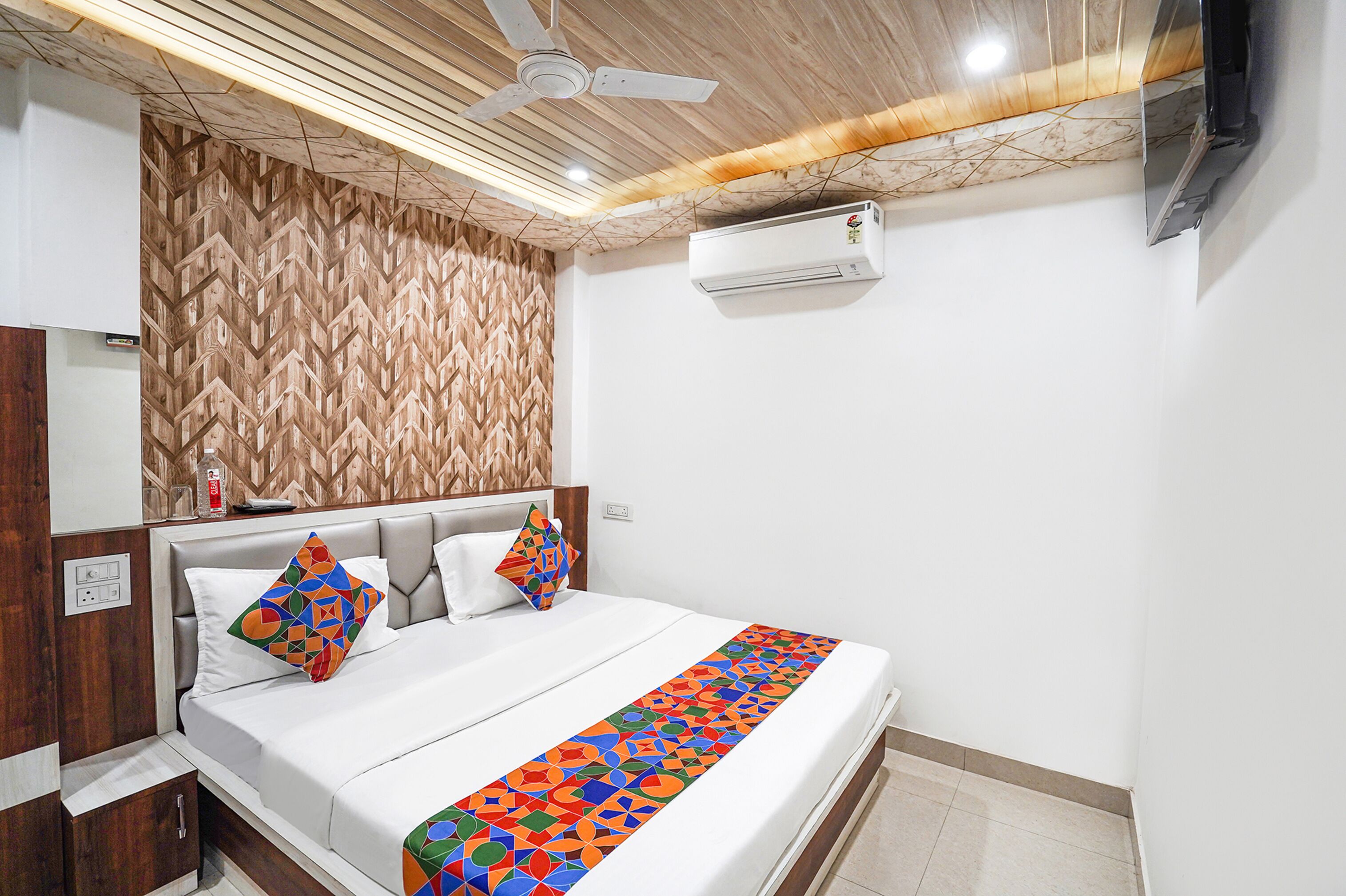 A hotel room with a bed, a ceiling fan, an air conditioning unit, and a wooden-patterned wall.