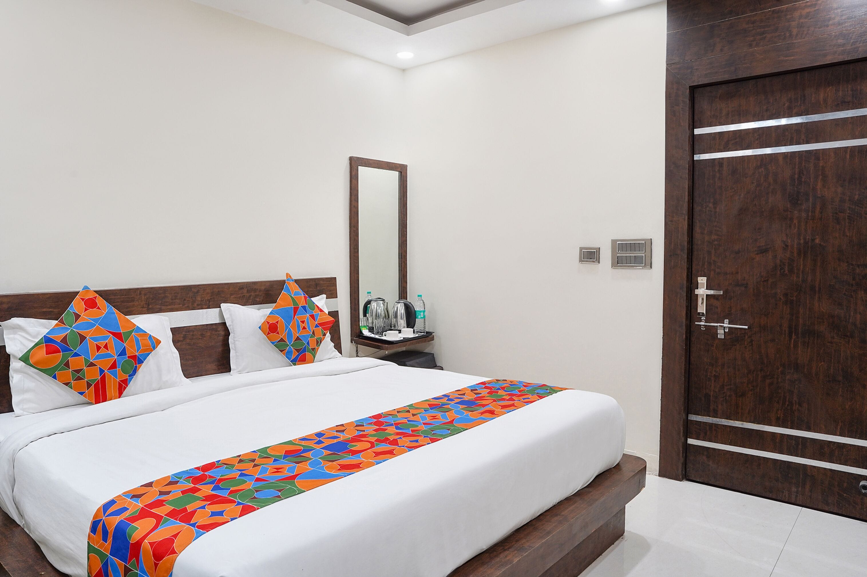 A hotel room with a neatly made bed, a wooden headboard, a large mirror, a wooden wardrobe, and a small table with toiletries.