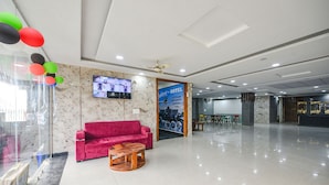 A hotel lobby with a red sofa, a wooden coffee table, a TV mounted on the wall, and a reception desk. - Fabhotel Radiant Blu (Noida)