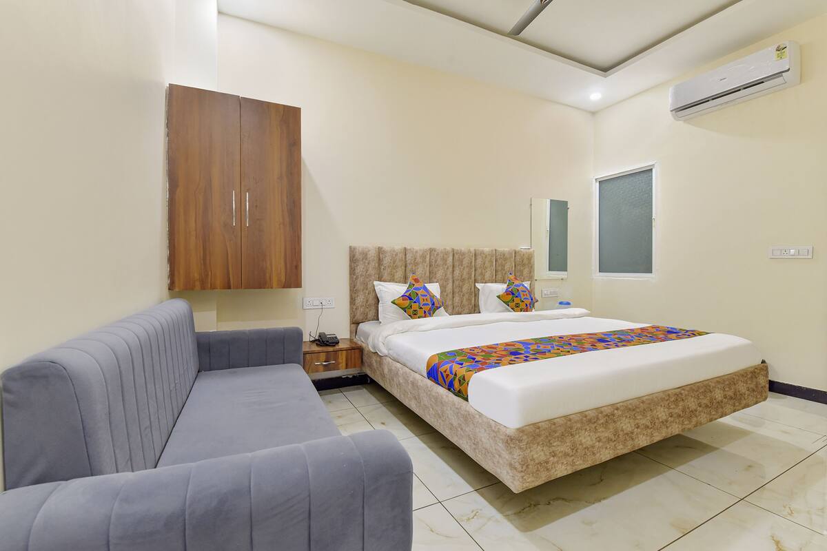 A bedroom with a bed, a sofa, a wooden wardrobe, a bedside table, and an air conditioning unit.