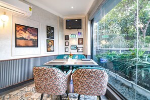 A dining area with a large table, two patterned chairs, framed pictures on the wall, and a view of greenery through a glass window.