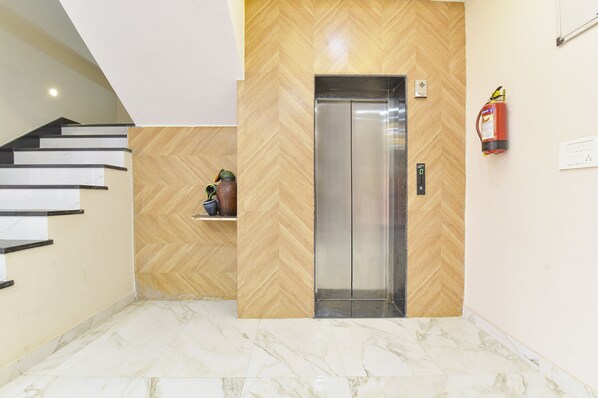 Un ascensor moderno con paneles de madera en diseño herringbone, suelo de baldosas de mármol y un extintor de incendios en la pared.