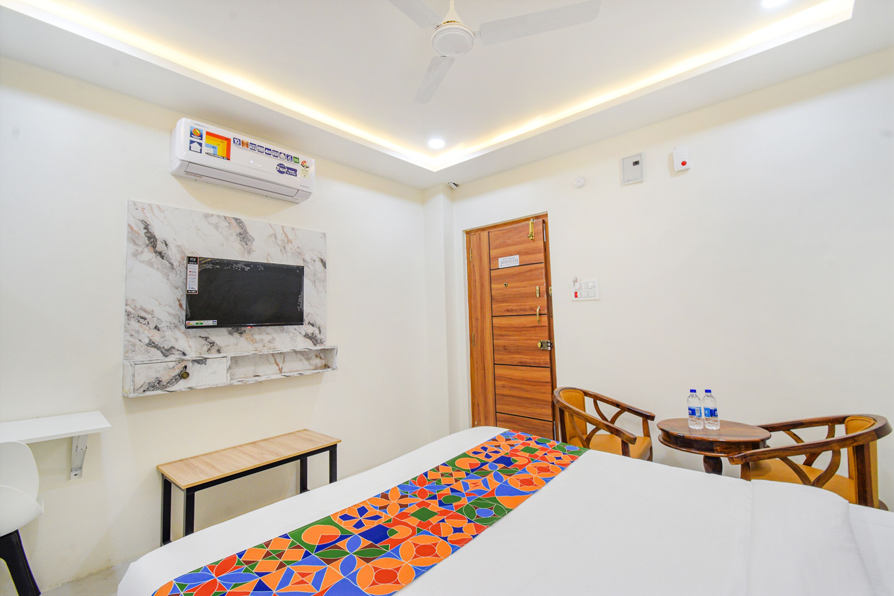 Habitación de hotel con cama, una mesita, una silla, televisor montado en una pared de mármol y una puerta de madera.