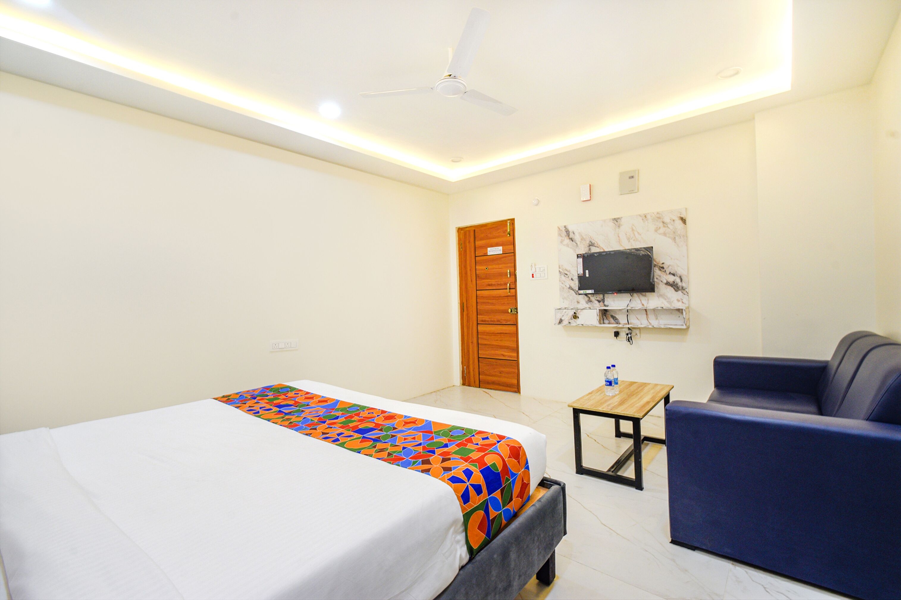 Una habitación de hotel con una cama, un sofá azul, una mesita, un ventilador de techo y un televisor montado en la pared.
