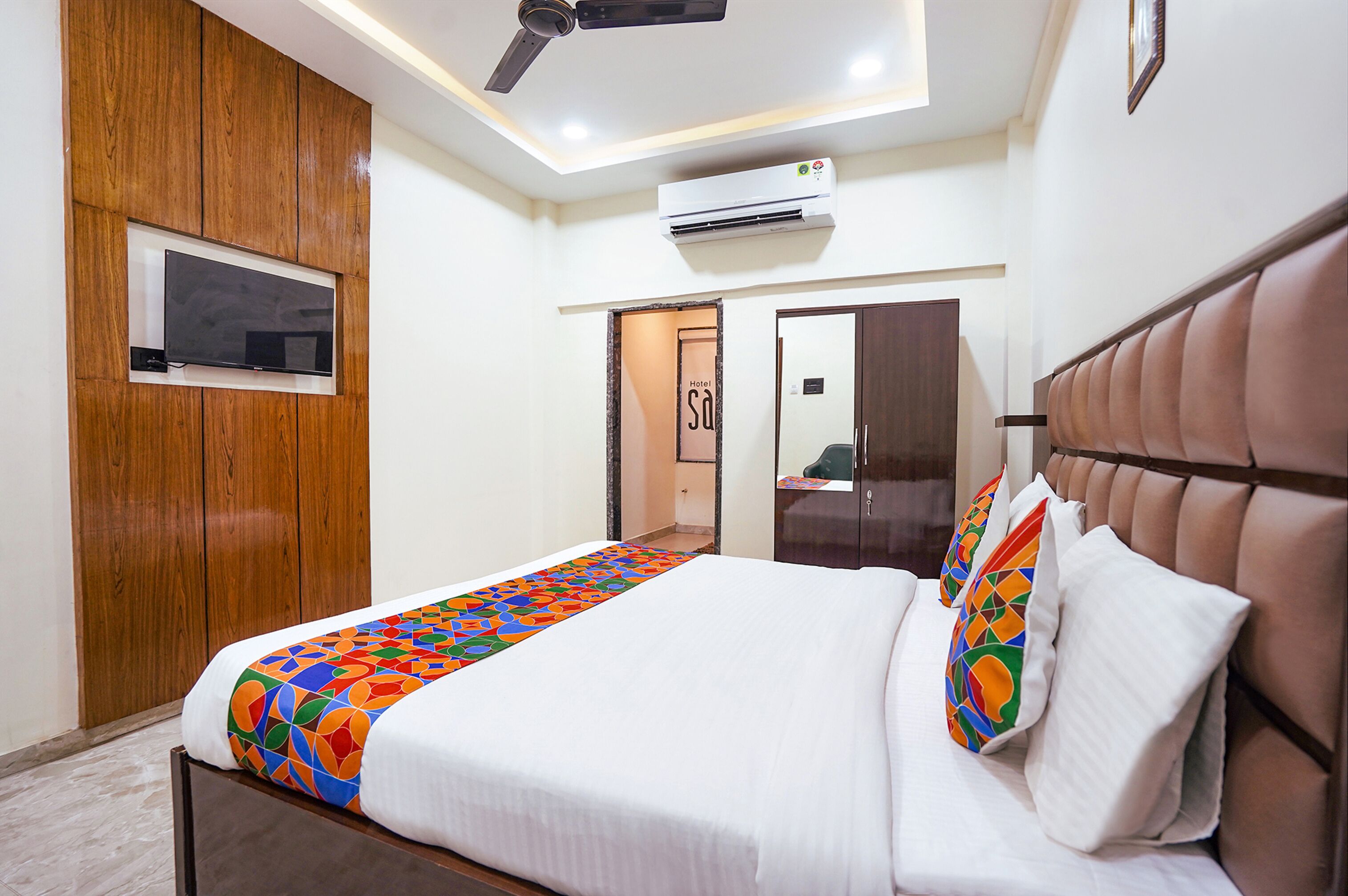 A hotel room with a bed, a ceiling fan, an air conditioning unit, a TV mounted on a wooden panel, and a door with a sign.