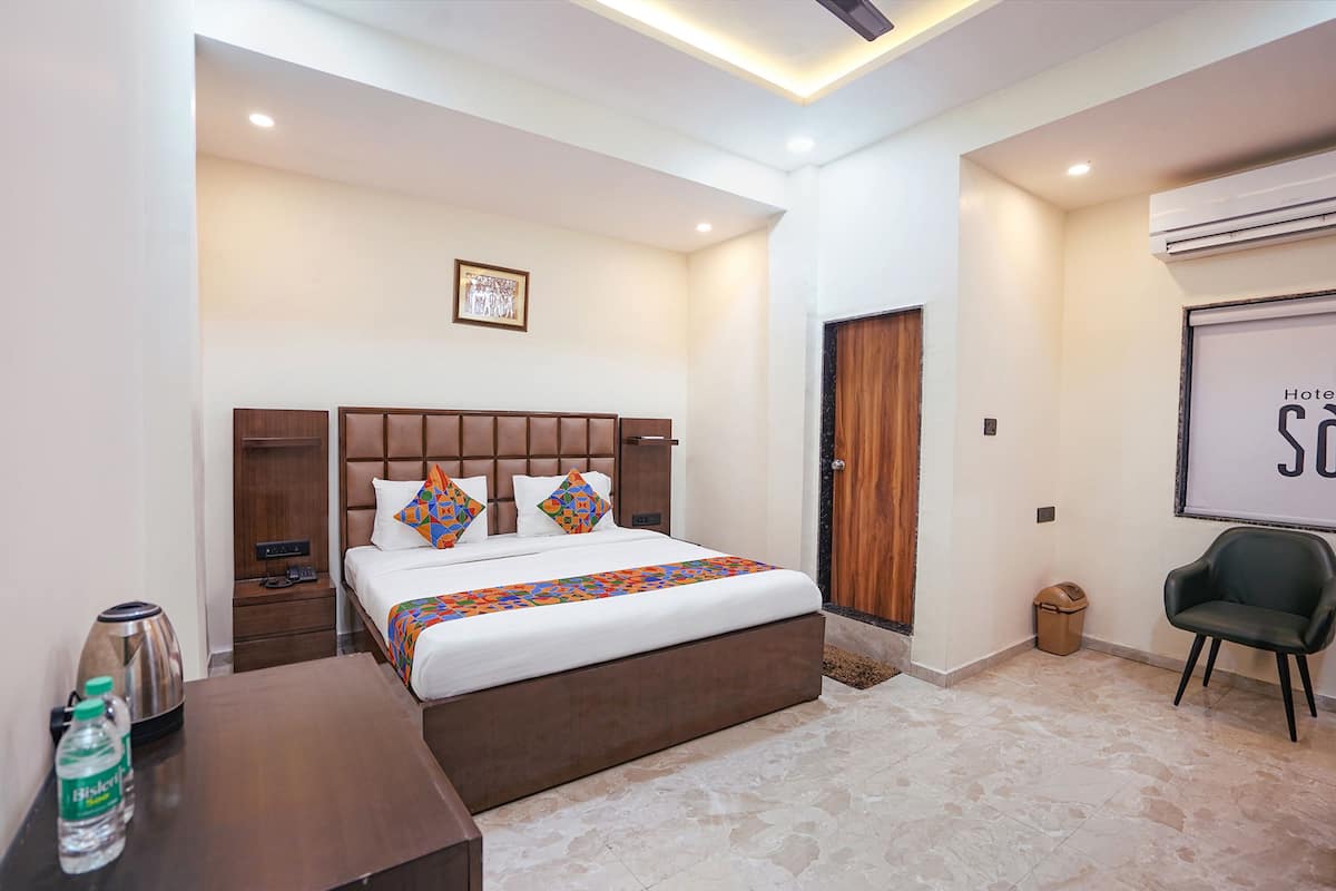 Una habitación de hotel con una cama grande, un cabecero de madera, una mesita de noche con hervidor y botella de agua, un cubo de basura y una silla.